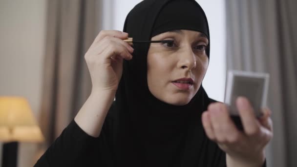 Närbild av ung vacker muslimsk dam tittar på liten spegel och tillämpa mascara. Modern österländsk kvinna i svart hijab sminkar sig. Skönhet, kosmetika. — Stockvideo