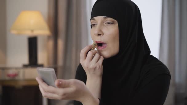 Porträtt av ung vacker muslimsk dam tittar på spegel och tillämpa läppstift. Modern kvinna i svart traditionell hijab gör make-up. Skönhet, kosmetika. — Stockvideo