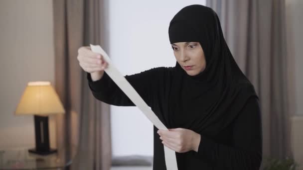 Jonge gestresste moslimvrouw in traditionele hijab verhogen wetsvoorstel en kijken naar het met geschokte gezichtsuitdrukking. Jonge oosterse dame die thuis met bonnetjes omgaat. Lifestyle, moslimcultuur. — Stockvideo