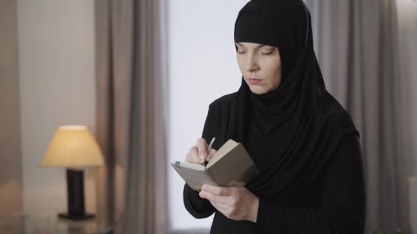 Portret van een bedachtzame moslimvrouw in hijab die schrijft in een notitieboekje. Een ijverige oosterse dame die een lijst van aankopen doet. Moslims, tradities, cultuur. — Stockvideo