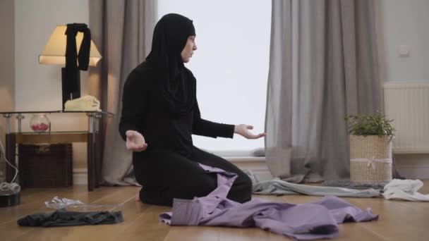Porträtt av desperat muslimsk kvinna sitter på golvet och tittar runt på utspridda kläder. Östra hemmafrun gör hushållsarbete. traditionell kultur, hushållning. — Stockvideo