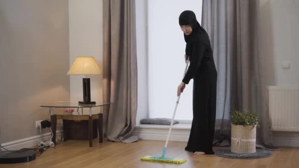 Mulher muçulmana sorrindo limpando o chão com esfregona. Jovem senhora em hijab e vestido tradicional fazendo tarefas domésticas. Serviço de limpeza, pessoas do leste . — Vídeo de Stock