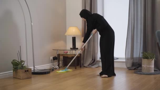 Porträtt av ung muslimsk dam som städar golvet. Ung dam i svart hijab använder mopp för hushållsarbete. Österländsk kultur, hushållning. — Stockvideo