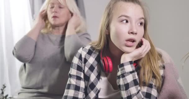 Teenager kaukasisches Mädchen sitzt mit irritiertem Gesichtsausdruck, während ihre reife Großmutter im Hintergrund den Kopf schüttelt. Kommunikationsprobleme, familiäre Beziehungen. kino 4k prores hq. — Stockvideo