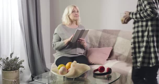 Portrait of senior blond Caucasian woman sitting on couch with tablet as her granddaughter coming up with coffee cups. Happy grandmother spending weekends with grandchild at home. Cinema 4k ProRes HQ. — Stock Video