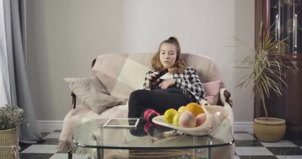 Retrato de una adolescente caucásica aburrida sentada en un sofá y cambiando de canal. Adolescente lindo viendo la televisión en casa los fines de semana. Adolescencia, estilo de vida, edad de transición. Sede del cine 4k ProRes . — Vídeo de stock