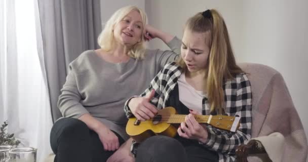 Portrait of teenage Caucasian girl playing ukulele for grandmother at home. Talented granddaughter entertaining mature woman on weekends. Cinema 4k ProRes HQ. — 비디오