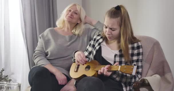 Young pretty Caucasian girl playing ukulele and singing for grandmother indoors. Talented granddaughter sitting with mature woman on couch. Cinema 4k ProRes HQ. — 비디오