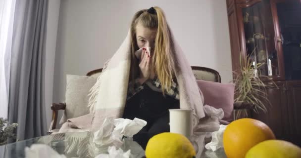 Πορτρέτο της αναστατωμένης καυκάσιας κοπέλας που φτερνίζεται και πίνει ζεστό τσάι. Πορτοκάλια και χρησιμοποιημένες χαρτοπετσέτες σκορπισμένες στο τραπέζι μπροστά σε έναν άρρωστο έφηβο. Πυρετός, καταρροή, αρρώστια. Κινηματογράφος 4k Prores Hq. — Αρχείο Βίντεο
