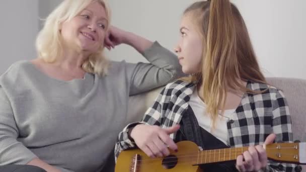 Portrait of cute young Caucasian girl playing ukulele and singing for grandmother at home. Talented granddaughter sitting with mature woman on couch indoors. Hobby, music. — Stock Video