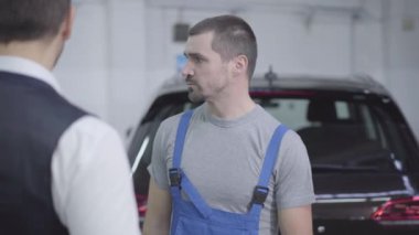 Kafkasyalı genç oto tamircisinin portresi tamirhanede müşteriyle konuşuyor. Kahverengi gözlü yakışıklı adam müşteriyle araba tamirini konuşuyor. Araba işi, tamir hizmetleri..