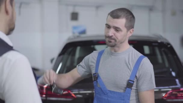 Retrato del joven trabajador caucásico que devuelve las llaves del coche al propietario después de la reparación de la garantía. Profesional mecánico de automóviles satisfecho con el resultado de su trabajo. Servicio, taller de reparación . — Vídeos de Stock