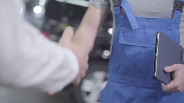 Fotocamera in movimento intorno alle mani di due uomini caucasici irriconoscibili in piedi in officina. Meccanico auto e cliente stringendo la mano sullo sfondo dei veicoli. Servizi di riparazione auto, servizi di riparazione . — Video Stock