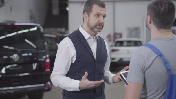 Cámara acercándose a la cara de cliente caucásico irritado hablando con mecánico de automóviles en taller de reparación. Hombre adulto guapo insatisfecho con el resultado del trabajo. Servicio, garantía . — Vídeos de Stock