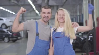 Beyaz erkek ve kadın güçlü bir hareket sergiliyor, kameraya bakıyor ve gülümsüyor. Tamirhanedeki atölyede çalışan profesyonel tamirci. Meslek, meslek, iş.