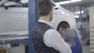 Yetişkin, beyaz bir adamın, araba tekerleklerinin vidalarını sıkan işçiye bakışını. Tamirhanedeki oto tamircisinin müşteri kontrolü. Hizmet, garanti.