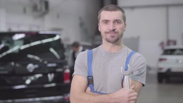 Portrét mladého pohledného bělocha, jak se dívá do kamery a usmívá se. Profesionální automechanik pózující na pracovišti s nastavitelným klíčem. Autoservis, záruka. — Stock video