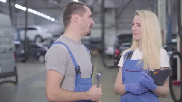 Retrato de dos mecánicos profesionales hablando, girando hacia la cámara y sonriendo. Hombre y mujer con túnicas azules de pie en el taller de reparación. Profesionales, ocupación, trabajo . — Vídeos de Stock