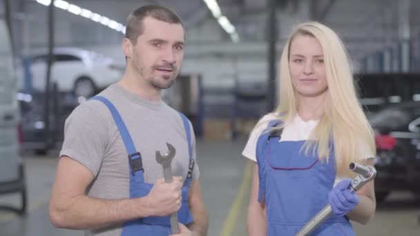 Giovani meccanici auto professionali guardando la fotocamera e sorridendo, parlando tra loro. Uomo e donna caucasici in abiti da lavoro in piedi sul posto di lavoro in officina di riparazione. Professione, occupazione, lavoro . — Video Stock