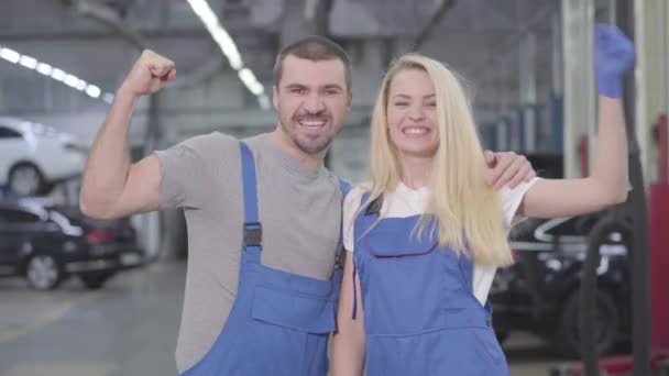Beyaz erkek ve kadın güçlü bir hareket sergiliyor, kameraya bakıyor ve gülümsüyor. Tamirhanedeki atölyede çalışan profesyonel tamirci. Meslek, meslek, iş. — Stok video