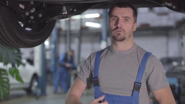 Porträt eines jungen ernsthaften kaukasischen Mannes in blauem Arbeitsmantel, der unter dem Auto in der Werkstatt steht und in die Kamera blickt. professioneller Automechaniker am Arbeitsplatz. Beruf, Beruf. — Stockvideo