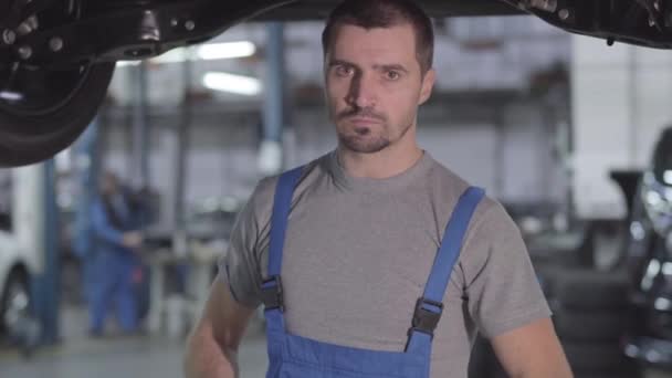 Kamera nähert sich einem ernsthaften kaukasischen Mann in blauer Robe, der unter dem Auto steht und in die Kamera schaut. professionelle junge Automechaniker in der Werkstatt. Beruf, Beruf. — Stockvideo