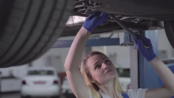 Camera moving from left to right around young Caucasian woman standing under car and tightening screws. Blond female auto mechanic working in repair shop. Lifestyle, occupation, profession. — 비디오