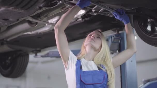 Camera approaching slowly to face of young Caucasian woman in workrobe standing under car and tightening screws. Confident female auto mechanic working in repair shop. Occupation, profession. — 비디오