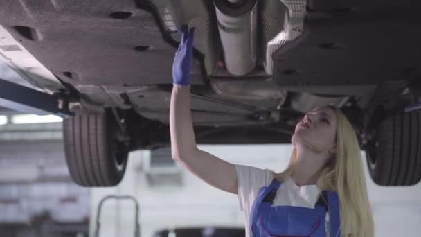 Onderaanzicht van zelfverzekerde blanke auto monteur controleren vaste auto. Blond jong vrouwtje dat in de garage werkt. Garantiedienst. — Stockvideo