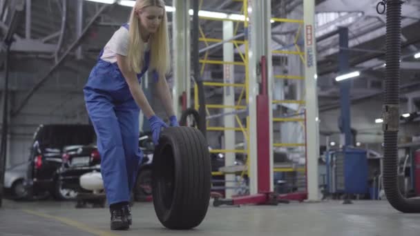 Jonge blanke vrouw die banden rolt in de garage. Lachende vrouwelijke automonteur op het werk. Camera uitzoomen. Levensstijl, werk, beroep. — Stockvideo