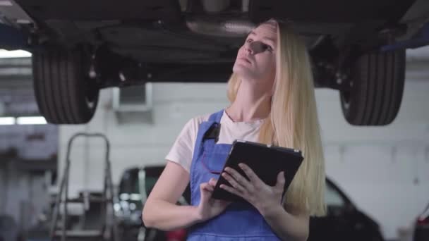 Portret van een jonge, mooie blanke vrouw in werkkleding die onder een kapotte auto staat met een tablet en fouten opmerkt. Vrouwelijke auto monteur vaststelling van auto. Professioneel, reparatiebedrijf. — Stockvideo
