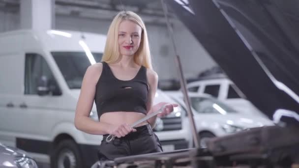 Porträtt av sexig blond vit kvinna som står bredvid öppen bilhuva med justerbar skiftnyckel och leende. Attraktiv bilmekaniker fastställande bil fel i reparationsverkstad. — Stockvideo