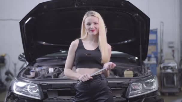 Sexy blond běloška stojící na pozadí otevřené kapoty s nastavitelným klíčem a s úsměvem. Krásná samice auto mechanik stojící v opravně. — Stock video