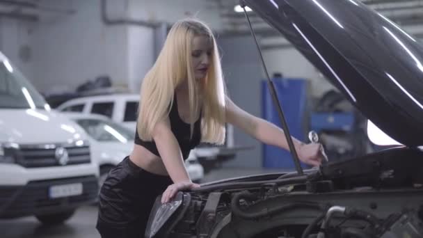 Attractive blond Caucasian woman tightening screws in open car hood. Sexy auto mechanic in black clothes working at repair shop. Profession, occupation, beauty. — 비디오