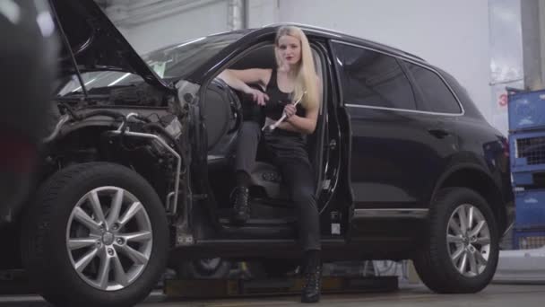 Porträt einer attraktiven kaukasischen blonden Frau, die mit verstellbarem Schraubenschlüssel am Fahrersitz sitzt und lächelt. schöner Automechaniker posiert in kaputtem Auto am Arbeitsplatz. — Stockvideo