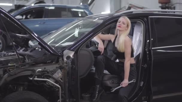 Atractiva mujer rubia caucásica posando en el asiento del conductor con llave ajustable. Hermoso mecánico de automóviles mirando a la cámara sentado en el automóvil roto en el lugar de trabajo . — Vídeo de stock