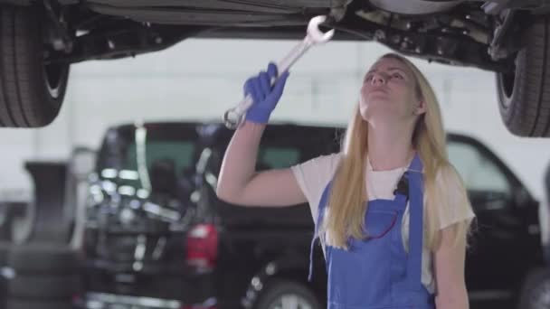 Retrato de la joven mujer caucásica de pie debajo del coche y tornillos de apriete. Mujer rubia mecánico de automóviles en bata de trabajo azul trabajando en taller de reparación. Estilo de vida, ocupación, profesión . — Vídeo de stock