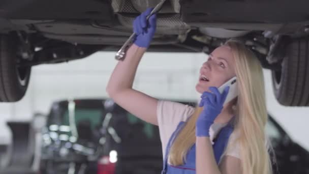 Ritratto di donna caucasica auto meccanico serraggio viti e parlare al telefono. Donna bionda in abito da lavoro blu in piedi sotto l'automobile in officina. Stile di vita, professione, professione . — Video Stock