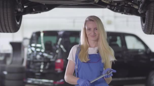 Portré fiatal női autószerelő munkaruhában áll az autó alatt a javítóműhelyben, nézi a kamerát és mosolyog. Magabiztos, fehér, szőke nő csavarkulccsal a kezében. Foglalkozás, munka, foglalkozás. — Stock videók