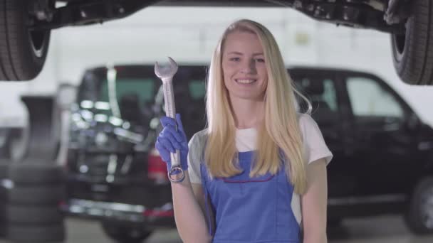 Glimlachende vrouwelijke automonteur in werkjas met moersleutel en duim omhoog. Blond blanke vrouw die naar de camera kijkt en glimlacht. Beroep, arbeid, beroep. — Stockvideo