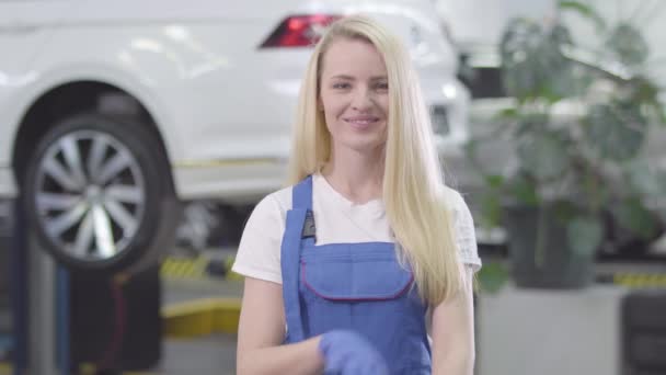 Portrait of young Caucasian woman standing at the background of white car in repair shop. Blond female auto mechanic crossing hands and smiling. Lifestyle, occupation, repair shop. — 비디오