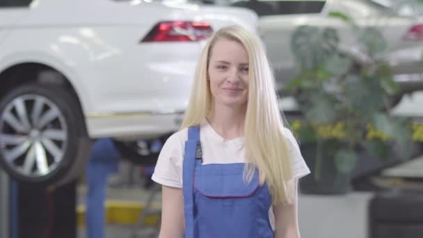 Vrolijke blanke vrouw die op de achtergrond van de auto staat en autosleutels van de camera laat zien. Blond vrouwelijke automonteur tevreden met haar werk. Lifestyle, beroep, motivatie. — Stockvideo
