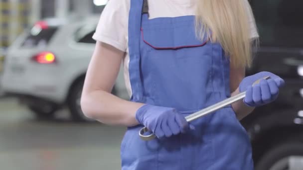 Unrecognizable blond woman in blue workrobe and gloves holding wrench. Young female auto mechanic standing at the background of car in repair shop. Lifestyle, occupation. — 비디오