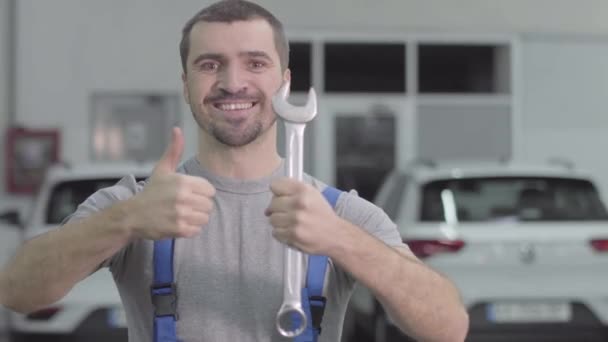 一个有着棕色眼睛的快乐的白人男子的画像，他的大拇指举着扳手。 快乐的男性汽车修理工看着相机，笑着站在修理店里。 生活方式、职业. — 图库视频影像