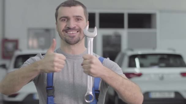 Portrait de l'homme blanc joyeux avec les yeux bruns montrant pouce vers le haut tenant clé. Joyeux mécanicien automobile masculin regardant la caméra et souriant comme debout dans l'atelier de réparation. Mode de vie, profession . — Video