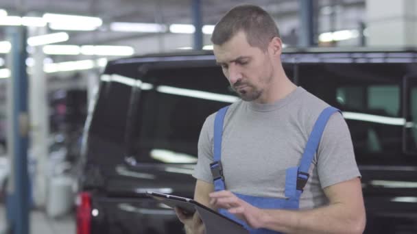 Mladý pohledný běloch v pracovním oděvu, stojící s tabletou na pozadí černého auta. Seriózní samec automechanik pracující v opravně. Sebevědomí, inspirace, životní styl. — Stock video