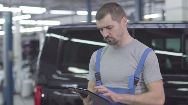 Joven hombre caucásico guapo en ropa de trabajo de pie con la tableta en el fondo del coche negro. Mecánico de automóviles masculino serio que trabaja en un taller de reparación. Confianza, inspiración, estilo de vida . — Vídeos de Stock