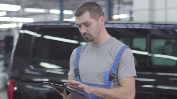 Portret van de jonge knappe blanke man in werkkleding met behulp van tablet, kijkend naar de camera en glimlachend. Zelfverzekerde vrolijke mannelijke automonteur op de achtergrond van de auto. Inspiratie, levensstijl, beroep. — Stockvideo