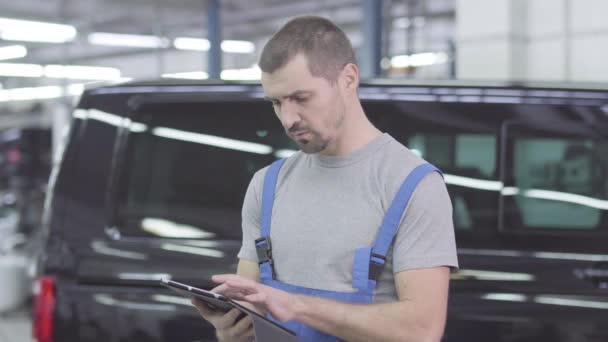 Portrét mladého bělošského automechanika, jak píše na tablet, dívá se do kamery a ukazuje palec nahoru. Radostný muž v dílně stojí na pracovišti v opravně. Záruka, servis. — Stock video