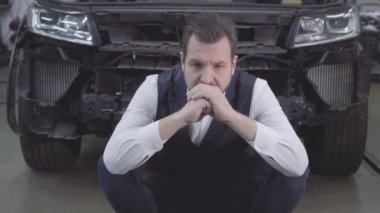 Çaresiz beyaz bir adamın portresi bozuk bir arabanın önünde oturuyor ve elleriyle başını tutuyor. Bozuk otomobilin arka planında üzgün bir yetişkin işadamı. Kaza, araba kazası, tamirhane..